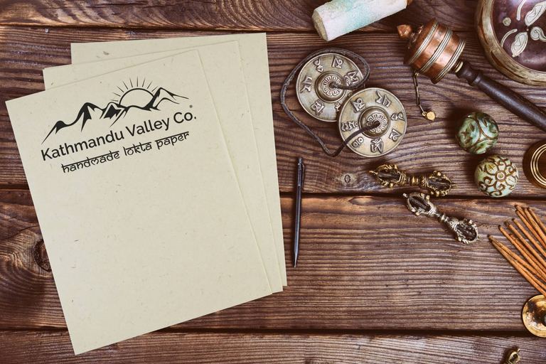 Sheets of Kathmandu Valley Co. handmade lokta printer paper on a wooden desk beside tingsha cymbals, a prayer wheel and a pen