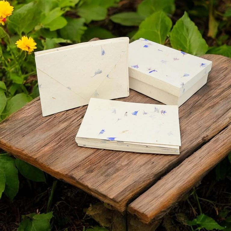 The Cornflower Cherish Card set on a weathered wooden step in a garden: a card, the envelopes and the petal-covered box