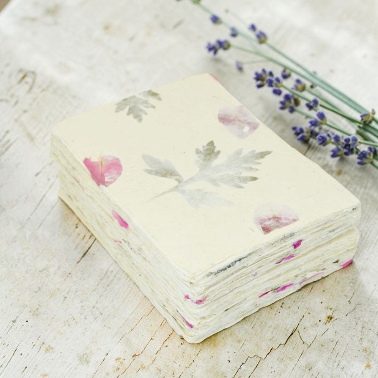 A stack of Forest Wanderer 5×7 card stock, bougainvillea petals and titepati leaves in the paper, on a white table with lavender