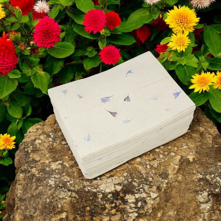 A stack of cornflower-petal 5×7 lokta envelopes on a rock among red and yellow garden flowers