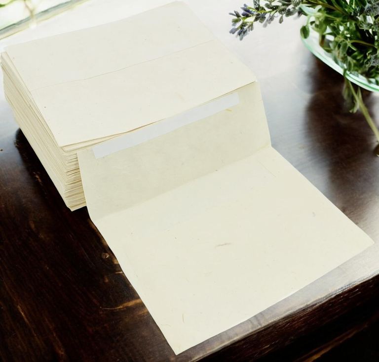 A natural lokta envelope, flap open, in front of a stack of them on a dark wooden table