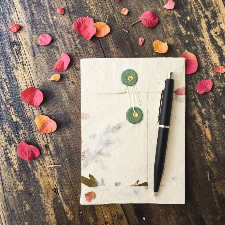 A Forest Wanderer pocket envelope with a pen laid on it for scale, bougainvillea petals scattered on dark wood