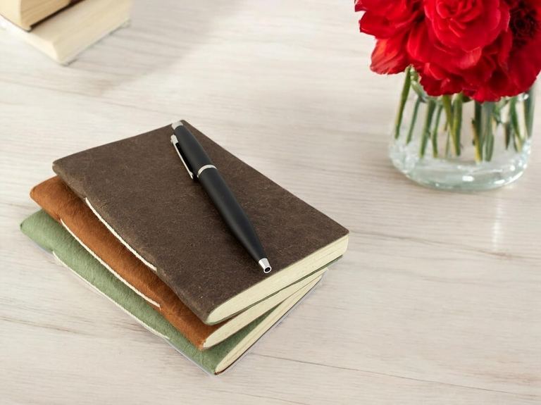 Three Nepali Companion notebooks in Dark Walnut, Terra Cotta and Sage stacked on a table beside red roses, a pen on top