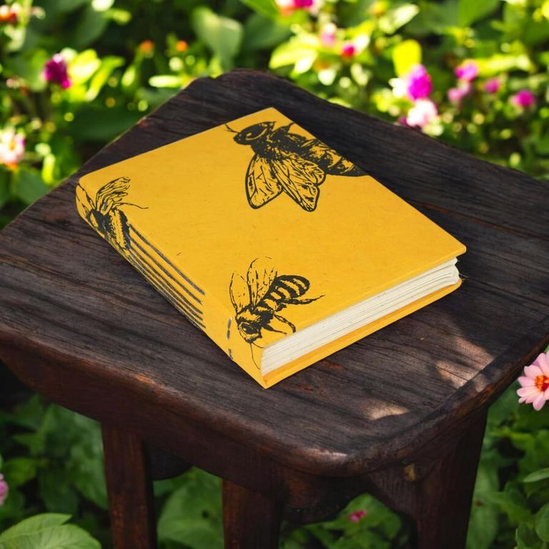 The small Busy Bee Nepali Keeper journal on a dark wooden stool among garden flowers
