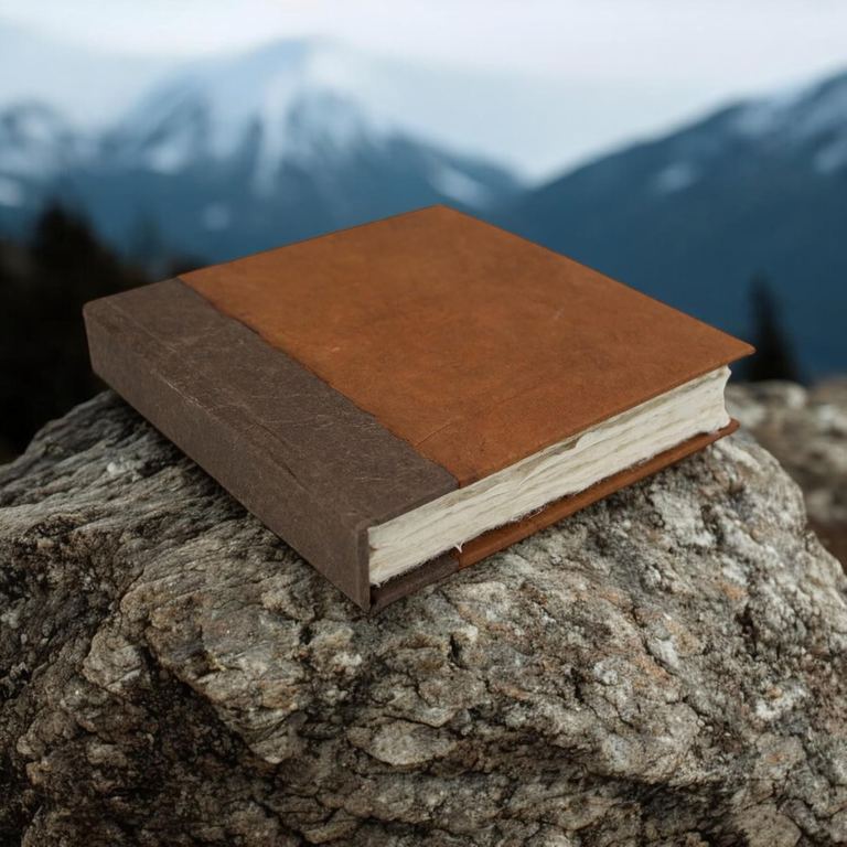 The 6 × 6 Terra Cotta Nepali Namaste journal on a lichen-covered rock with snowy mountains behind