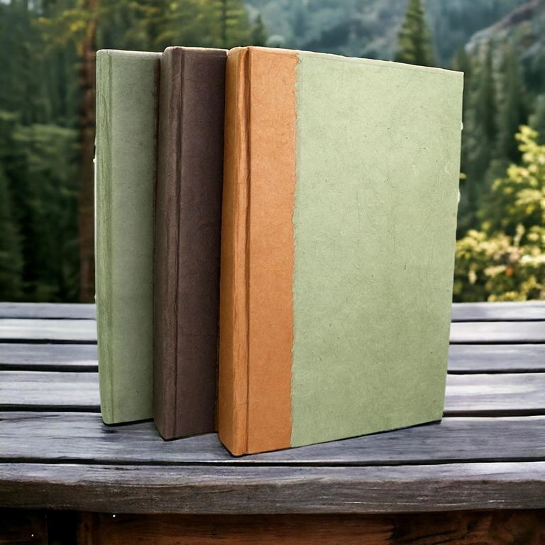 Sage, Dark Walnut and Terra Cotta Nepali Namaste journals standing together on a bench above a pine forest