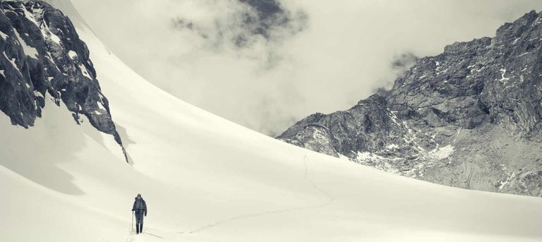 A lone trekker walking up a snowfield between rocky ridges, in black and white